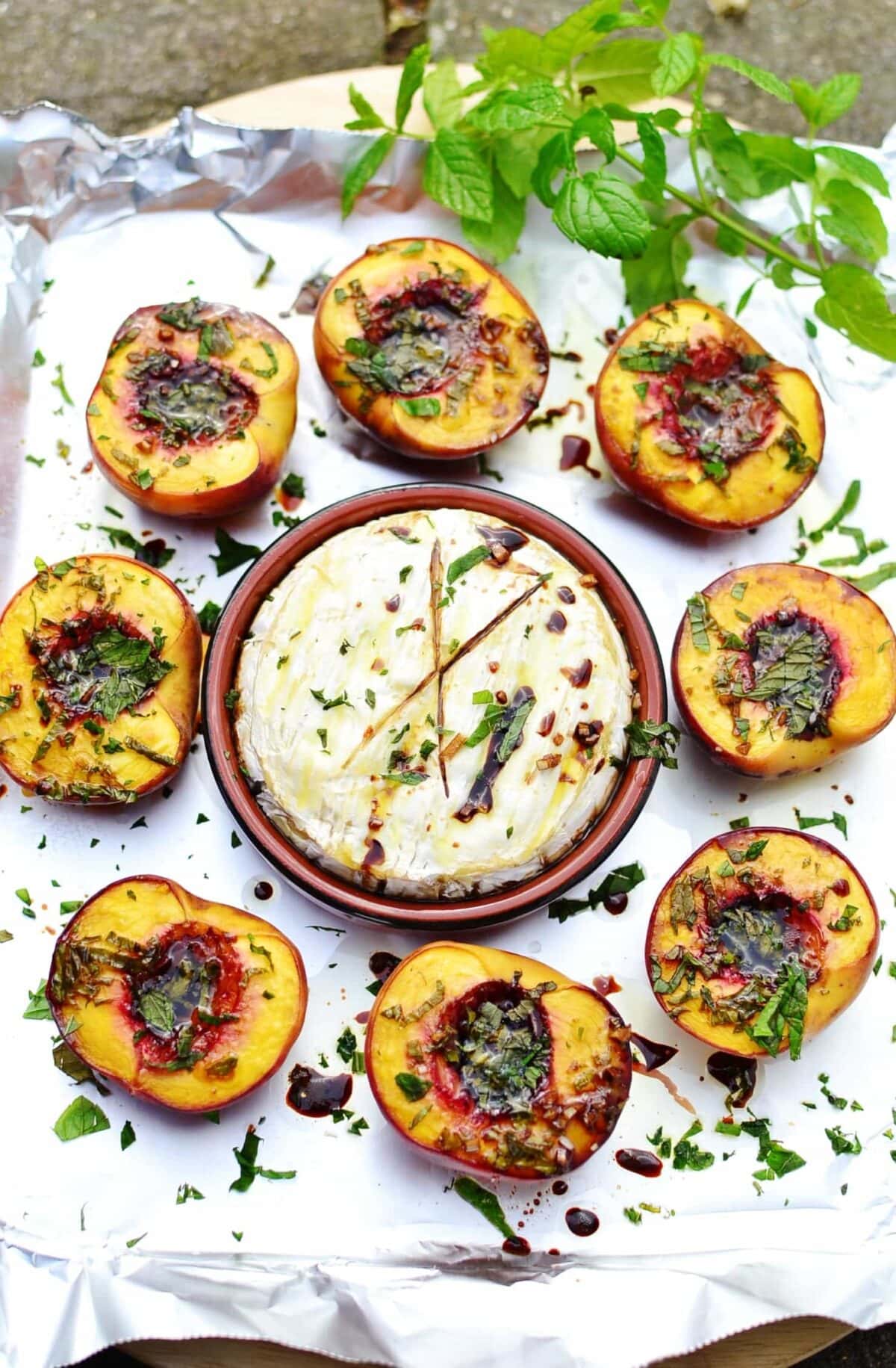 Top down view of baked camembert in round brown dish surrounded by baked halved peaches, with fresh mint.