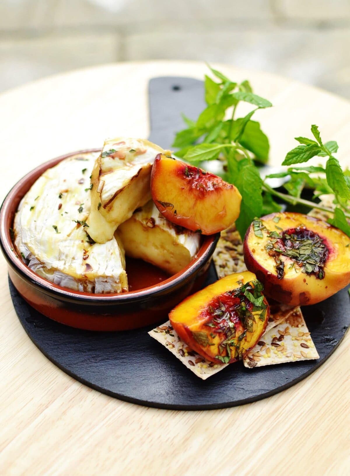 Baked camembert in round brown dish, with halved peaches, crackers and herbs on top of small slate platter.