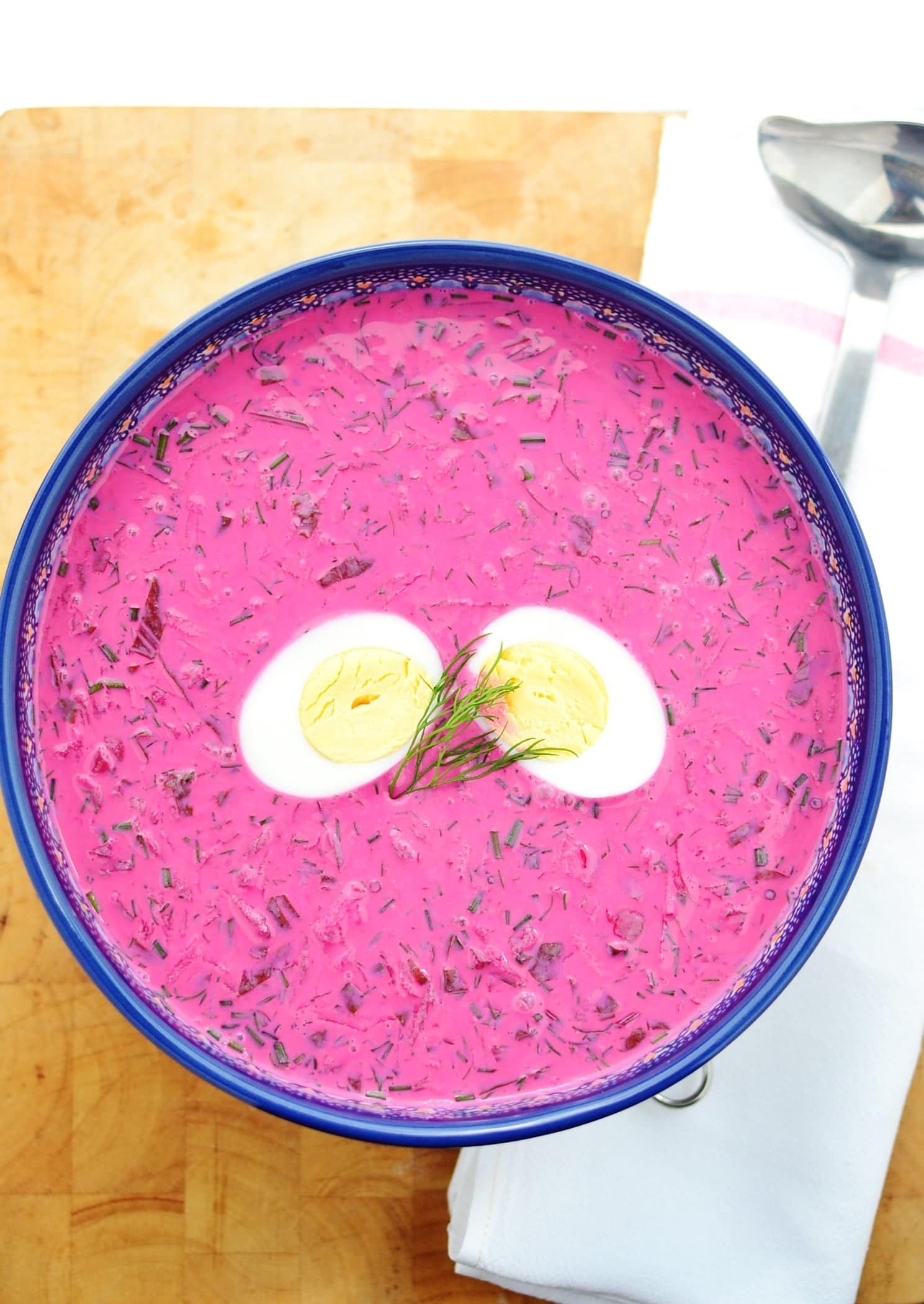 Top down view of cold beet soup with hard boiled egg halves in blue bowl with ladle on white cloth on top of wooden board.