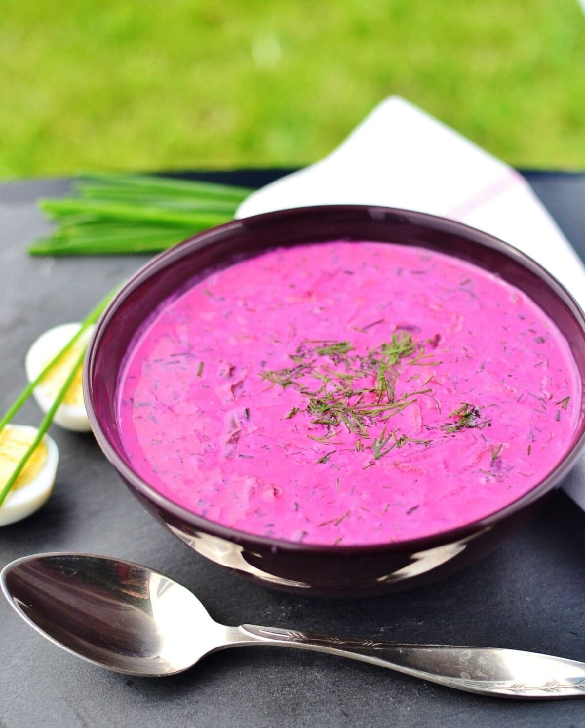 Cold beet soup in purple bowl with spoon in front, boiled eggs, chives and white cloth in background.