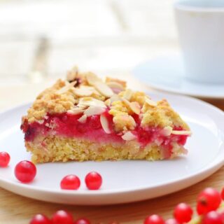 Side view of redcurrant cake slice on white plate with redcurrants on wooden surface.