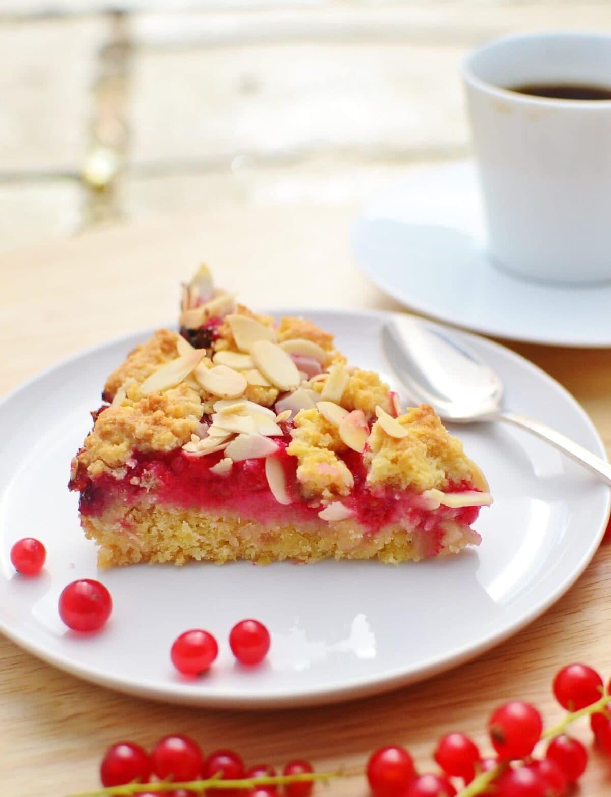 This simple Almond Redcurrant Cake recipe is a delicious way of enjoying seasonal fruit. It's tangy and sweet, low in fat as well as sugar and incredibly easy to make! #almondcake #redcurrantcake #redcurrants #healthydesserts #lowcarbcake #almondrecipe Side view of almond redcurrant cake slice on white plate with redcurrants on wooden surface with white cup and saucer in background.