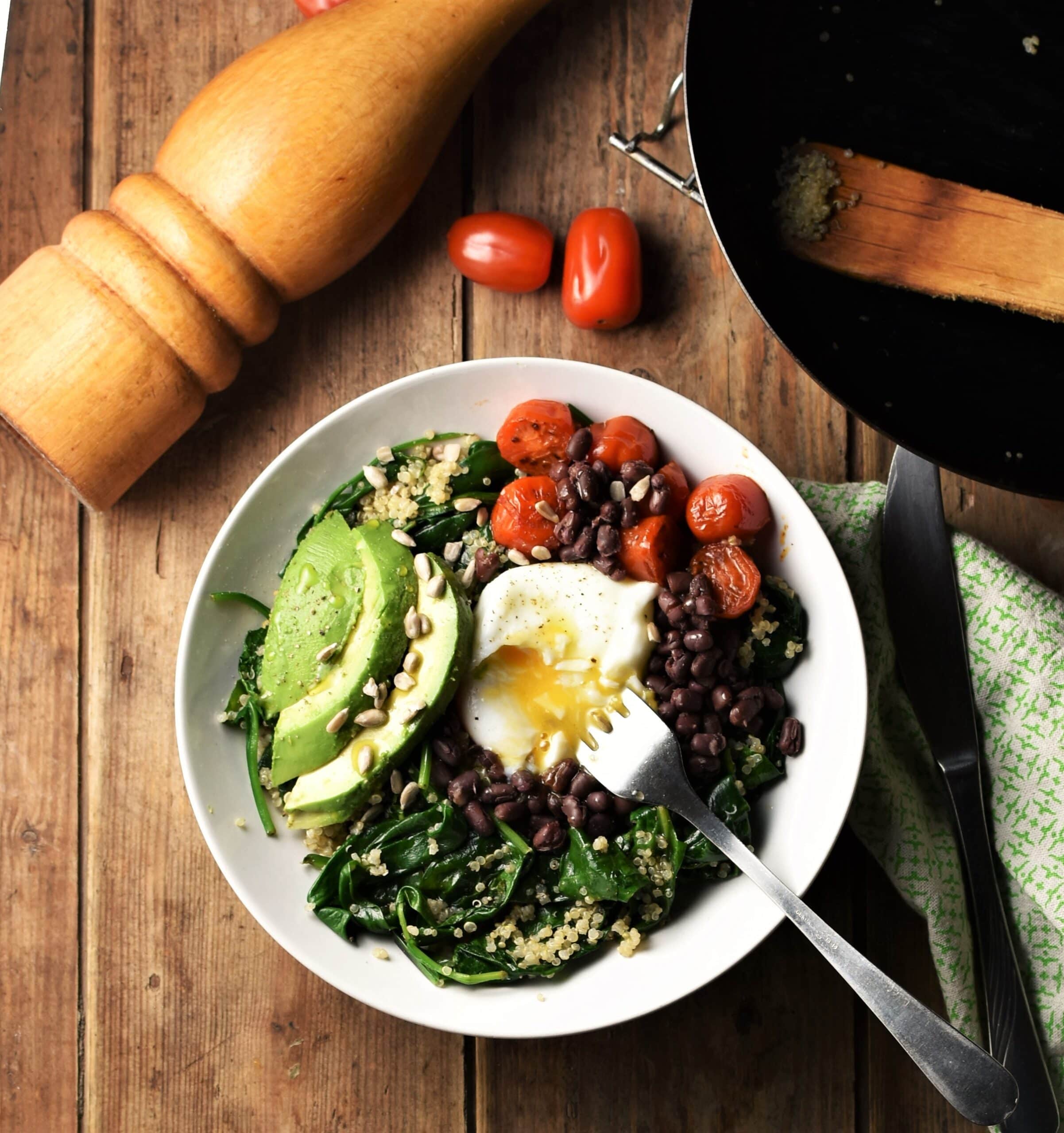 Quinoa and spinach mixture with avocado slices, cherry tomatoes, egg and beans in white bowl with fork, tomatoes, pepper grinder, green cloth with knife and skillet in background.