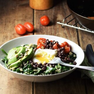 Side view of quinoa breakfast bowl with spinach, sliced avocado, egg, baby tomatoes and fork, with tomatoes, skillet and green cloth with knife in background.