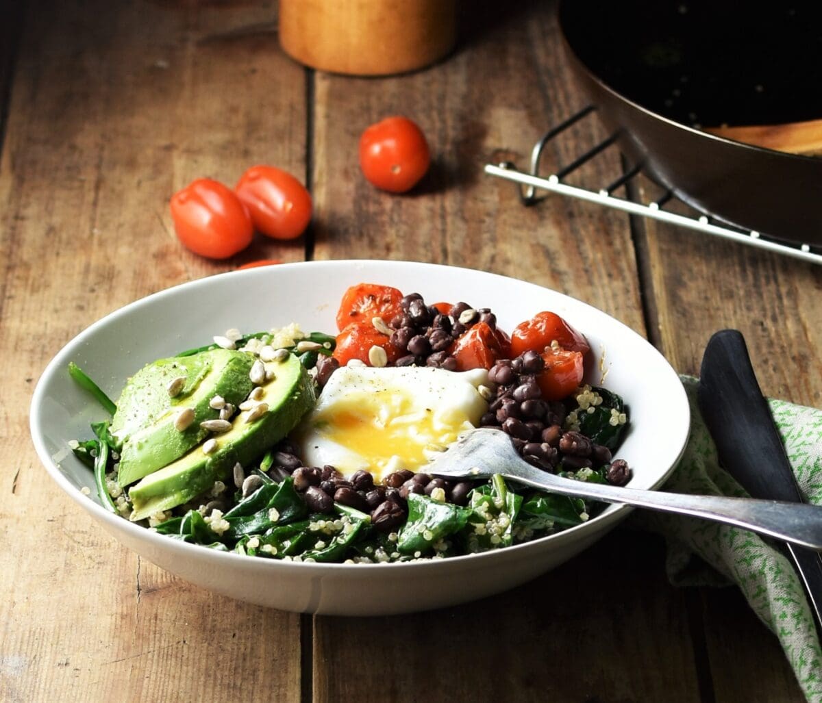 Side view of quinoa breakfast bowl with spinach, sliced avocado, egg, baby tomatoes and fork, with tomatoes, skillet and green cloth with knife in background.