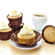 Cupcakes with white frosting in brown paper cases with saucer and white cup with coffee in background.
