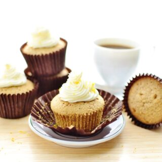 Cupcakes with white frosting in brown paper cases with saucer and white cup with coffee in background.