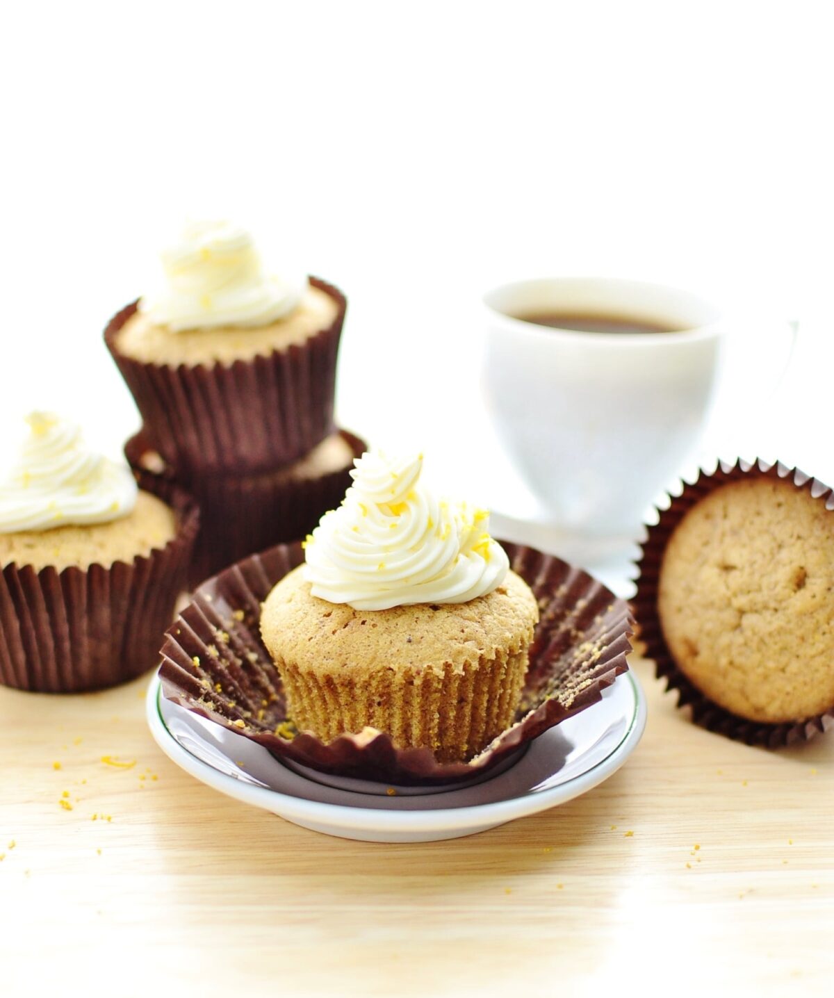 Cupcakes with white frosting in brown paper cases with saucer and white cup with coffee in background.