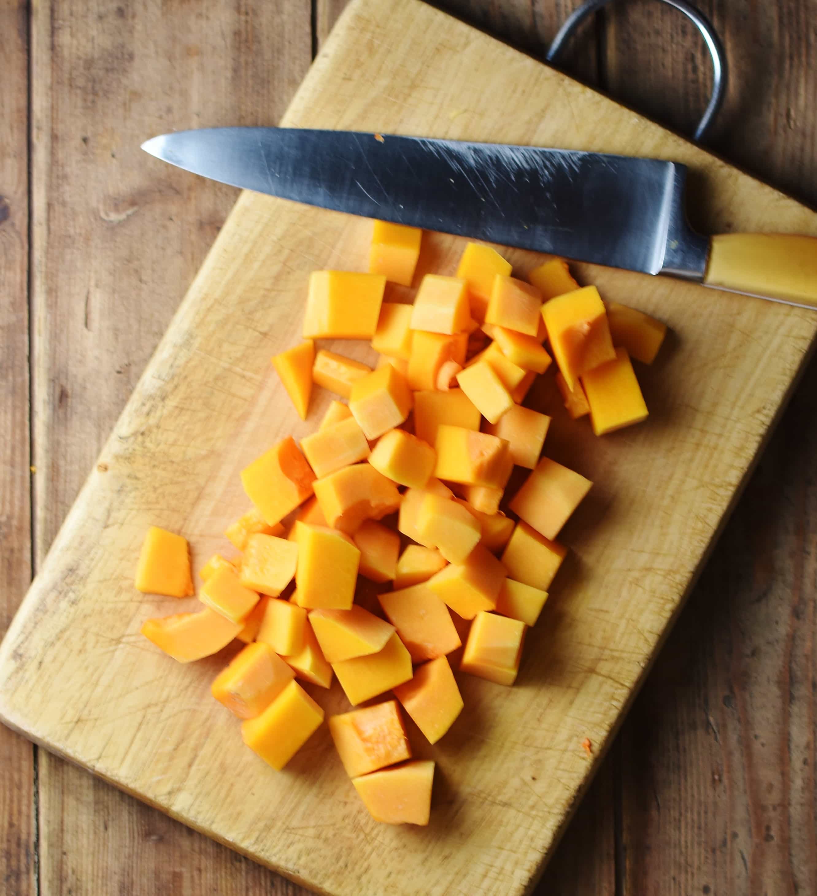 Peeled and cubed butternut squash on top of cutting board with knife.