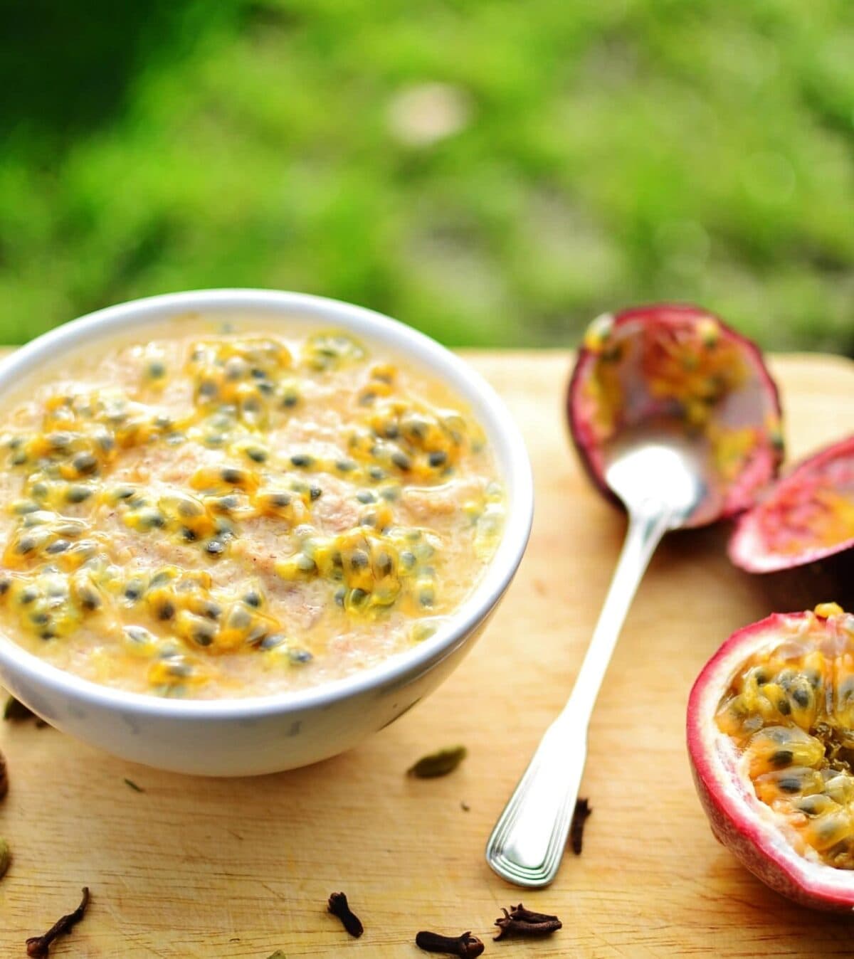 Chai spiced porridge with passion fruit in white bowl to left and spoon with cut passion fruit to right on light wooden surface with green background.