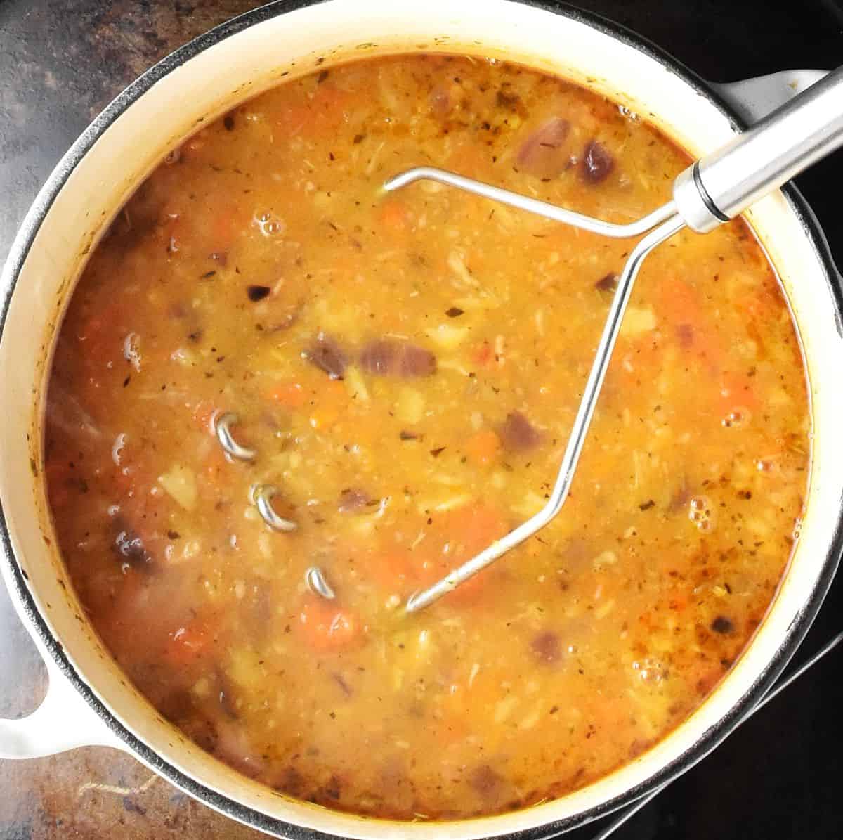 Thick chunky root vegetable soup in white pot with potato masher.
