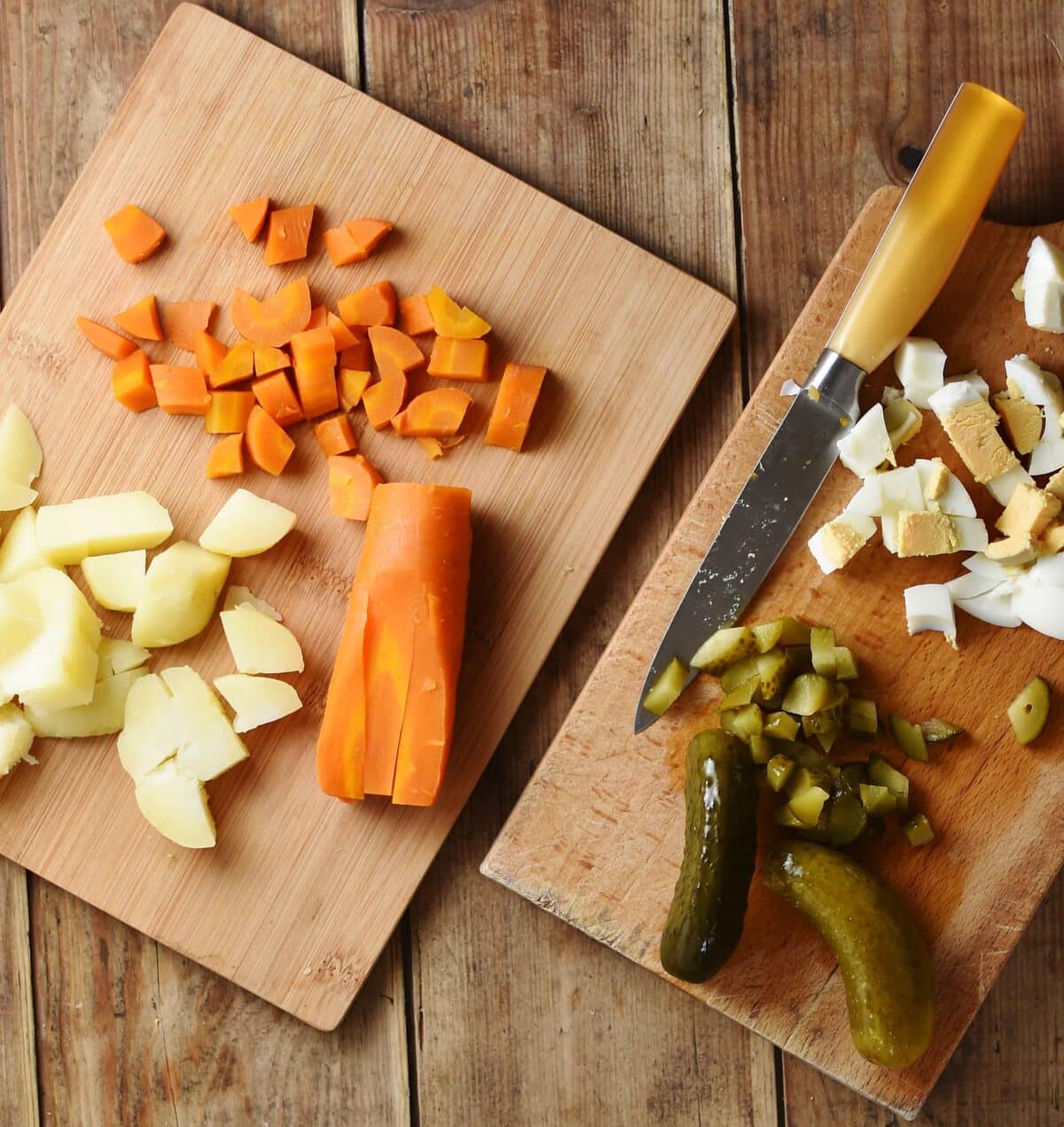 Top down view of chopped potatoes, carrots, eggs, gherkins and knife on top of 2 cutting boards.