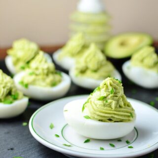 Deviled eggs with avocado on small white plate with garnish of chives and deviled eggs in background.