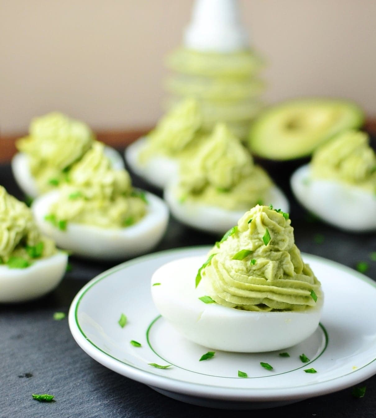 Deviled eggs with avocado on small white plate with garnish of chives and deviled eggs in background.