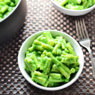 Top down view of spinach macaroni cheese in white bowl, with fork to right and more macaroni cheese in bowl and saucepan at top.