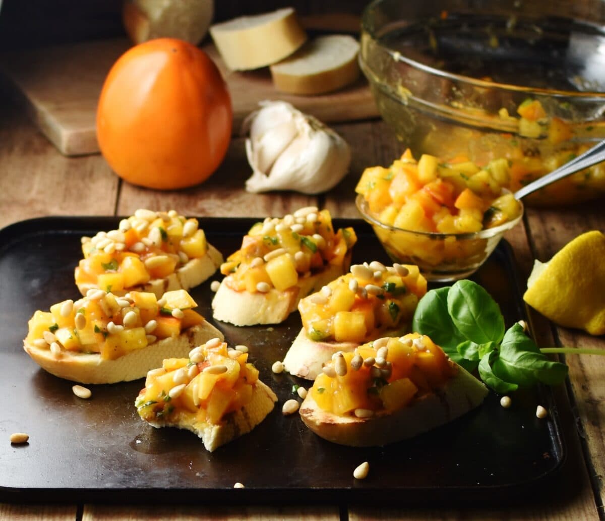 Persimmon mixture with pine nuts on top of baguette slices, with persimmon, garlic and mixing bowl in background.