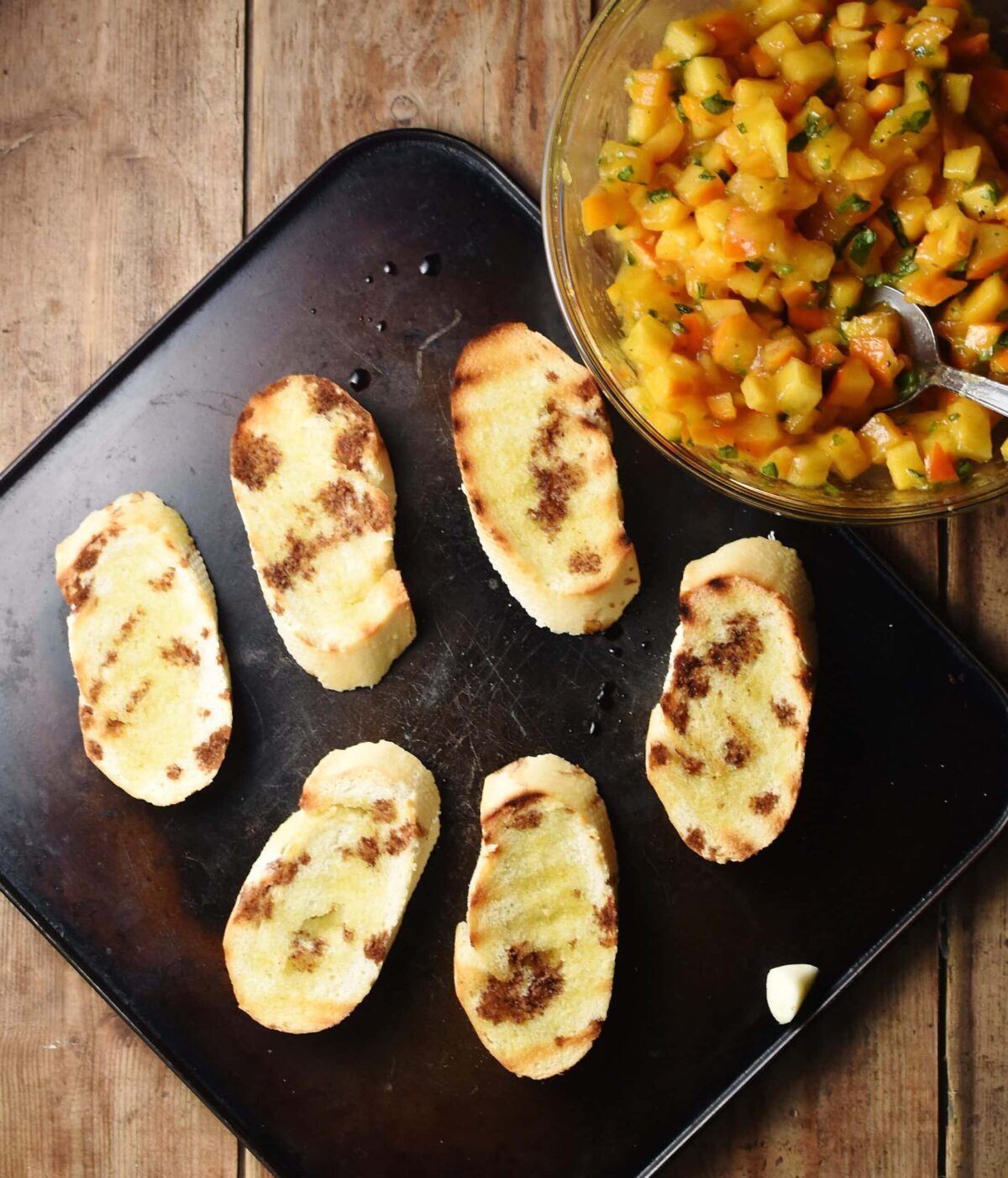 Grilled baguette slices drizzled over with balsamic vinegar on top of black tray, with chopped persimmon mixture in mixing bowl in top right.