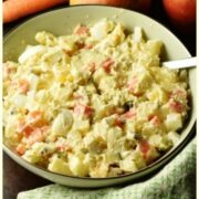 Close-up view of Polish potato salad with spoon in green bowl wrapped in green cloth, with carrot, apples and potatoes in background.