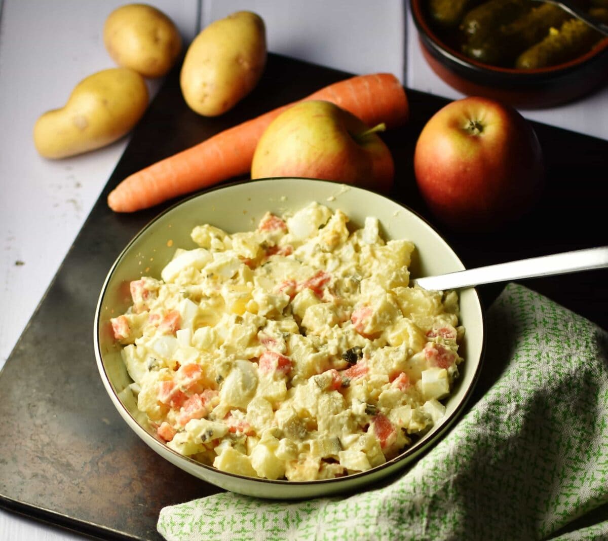 Potato salad with spoon in green bowl on top of oven tray, with green cloth in bottom right corner and raw carrot, apples and potatoes in background.