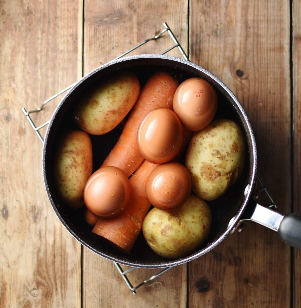 Top down view of unpeeled potatoes, carrots and eggs in large pot with water.