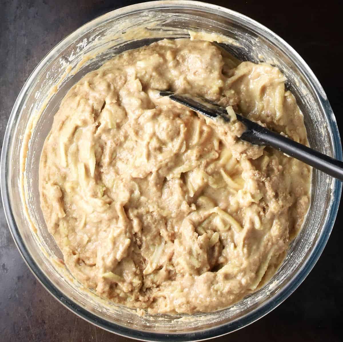 Top down view of gluten free muffin batter with grated apple in glass bowl with black spoon.