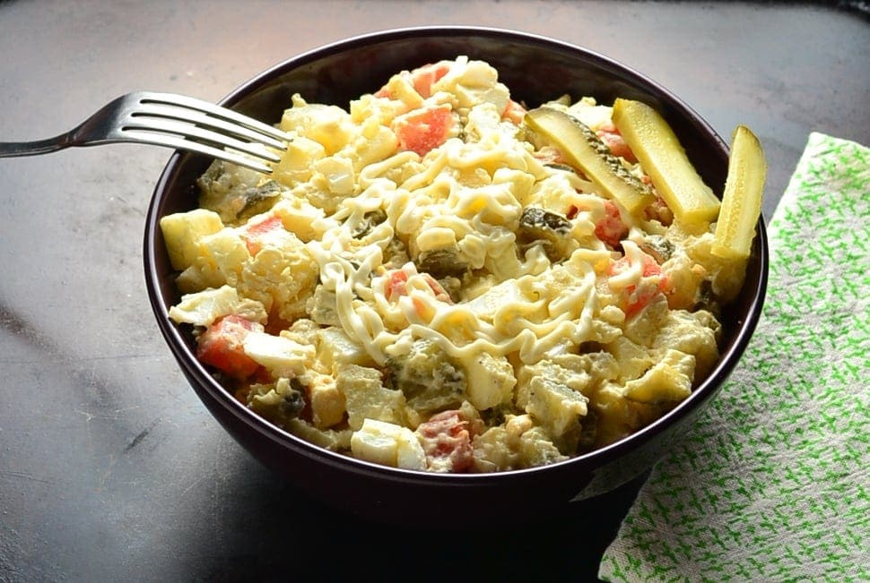 Polish potato salad in purple bowl, with fork on the left and green cloth on the right on dark brown surface.