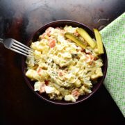 Top down view of Polish potato salad in purple bowl, with fork on the left and green cloth on the right on dark brown surface.