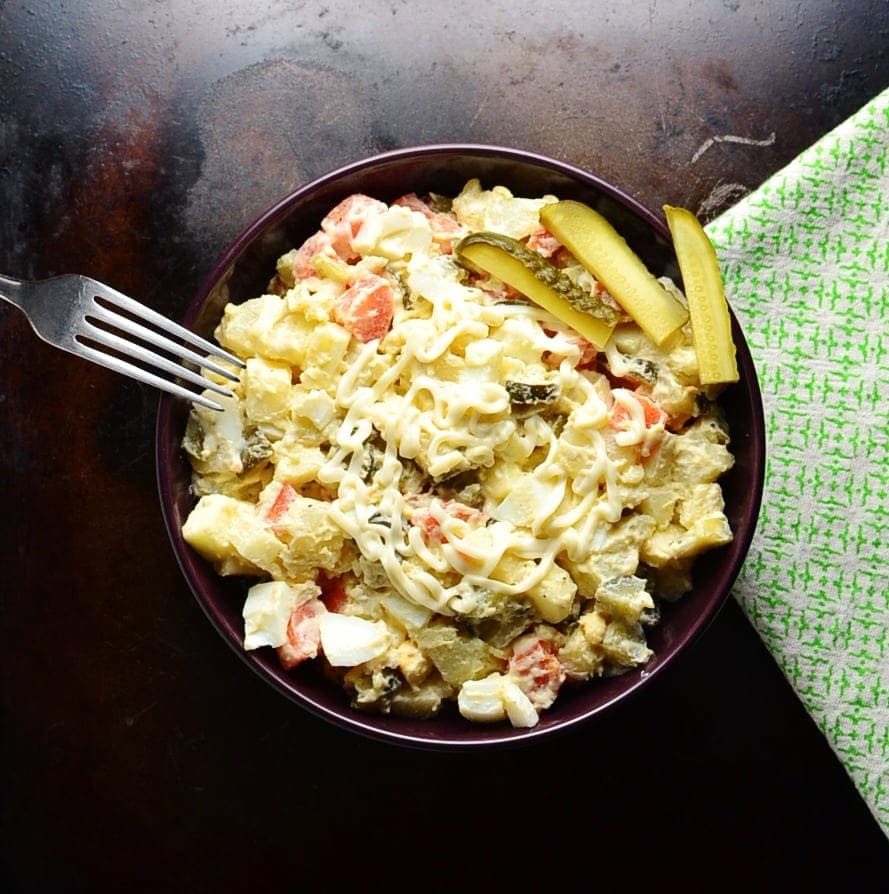 Top down view of Polish potato salad in purple bowl, with fork on the left and green cloth on the right on dark brown surface.