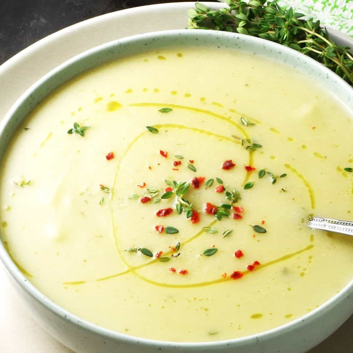 Close-up view of creamy potato leek cauliflower soup in green bowl with spoon and garnish of herbs.