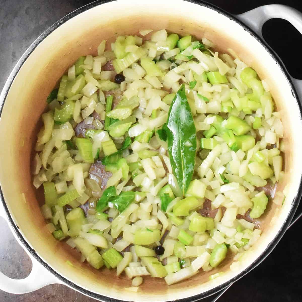 Soup base vegetable mixture with bay leaf in large white pot.