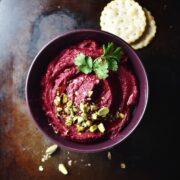 Beetroot dip with pistachios and herbs inside purple bowl, with crackers in background.