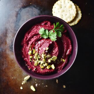 Beetroot dip with pistachios and herbs inside purple bowl, with crackers in background.