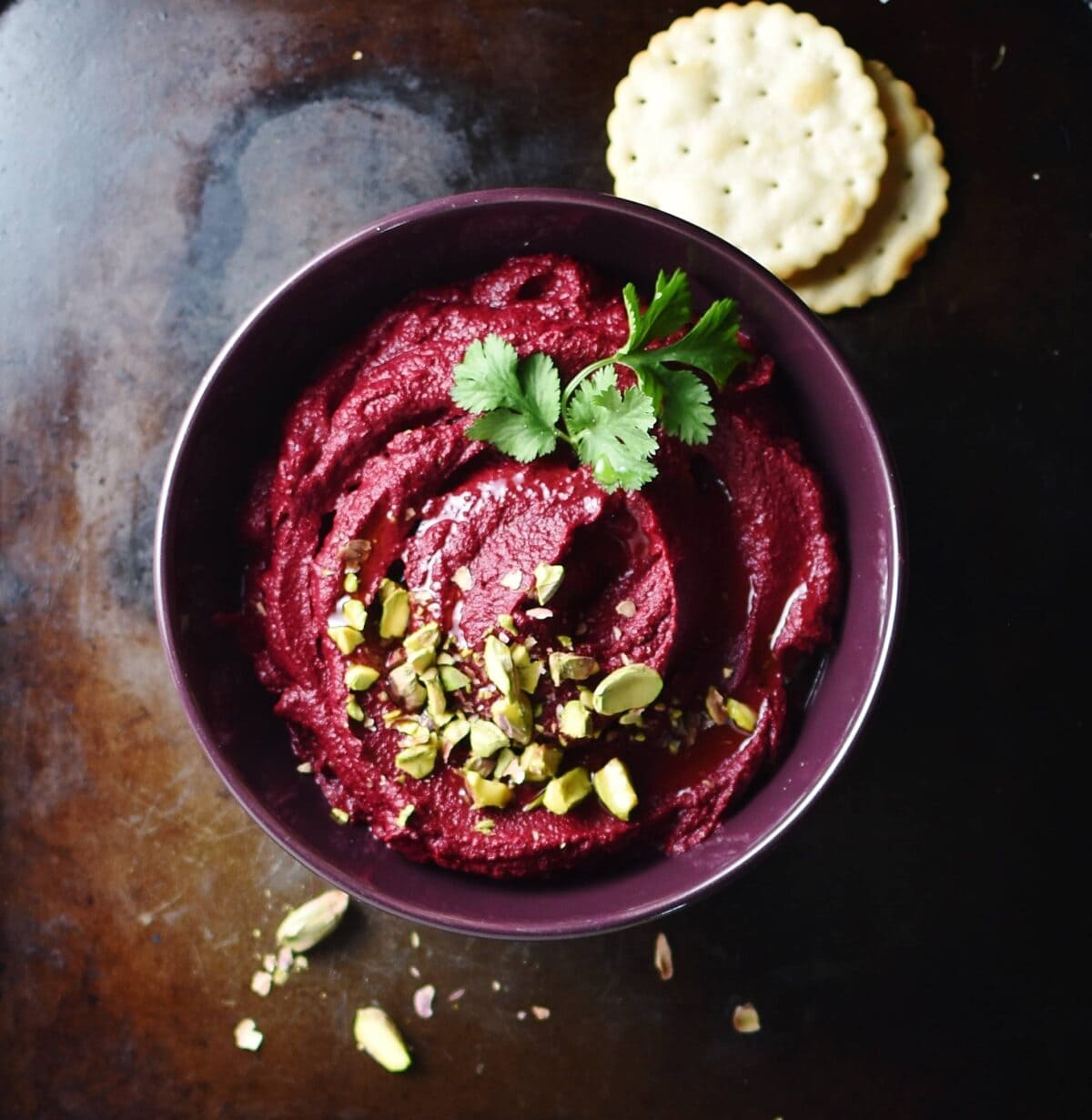 Beetroot dip with pistachios and herbs inside purple bowl, with crackers in background.