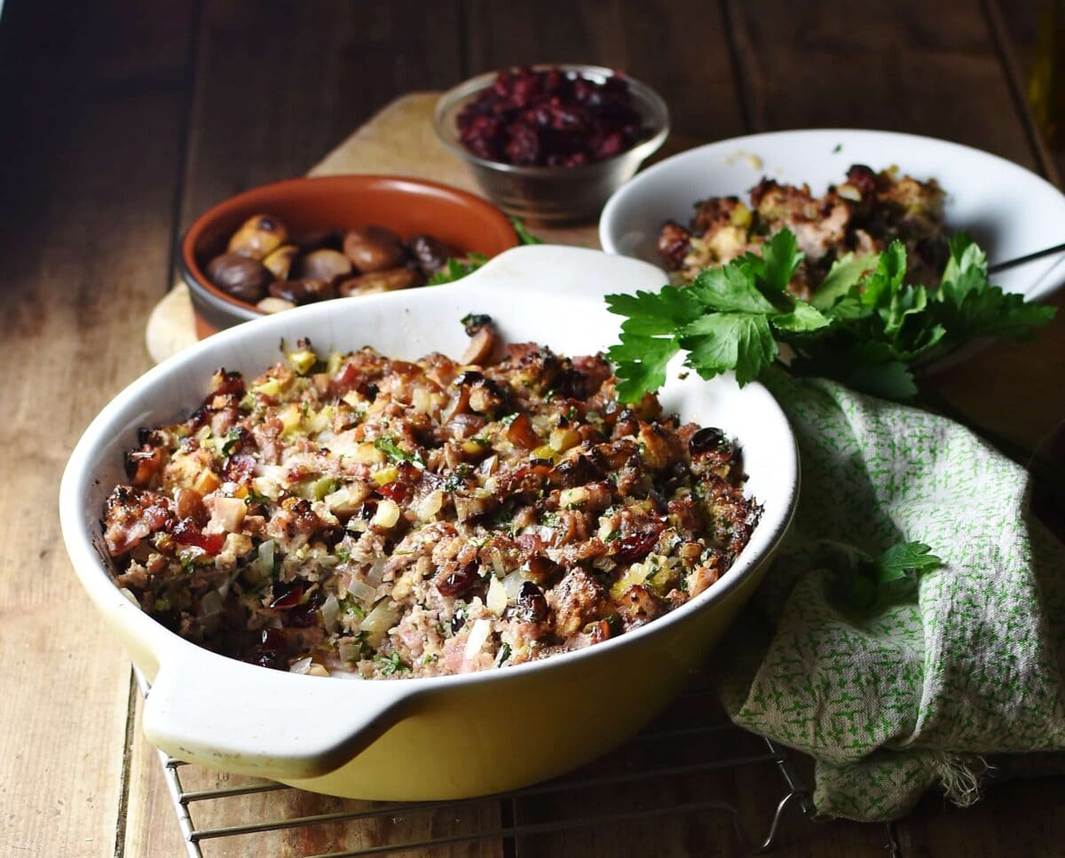 Side view of stuffing in white casserole dish, with green cloth, stuffing in white bowl and chestnuts in brown dish in background.