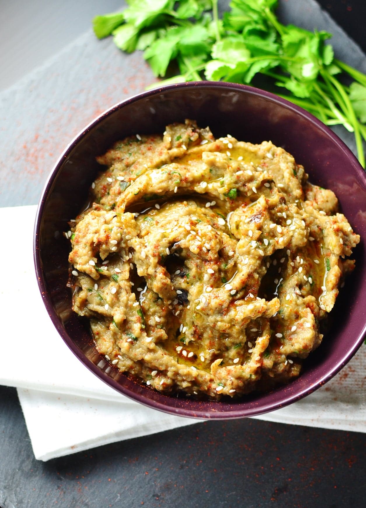 Top down view of eggplant dip in purple bowl on top of white cloth with fresh cilantro.