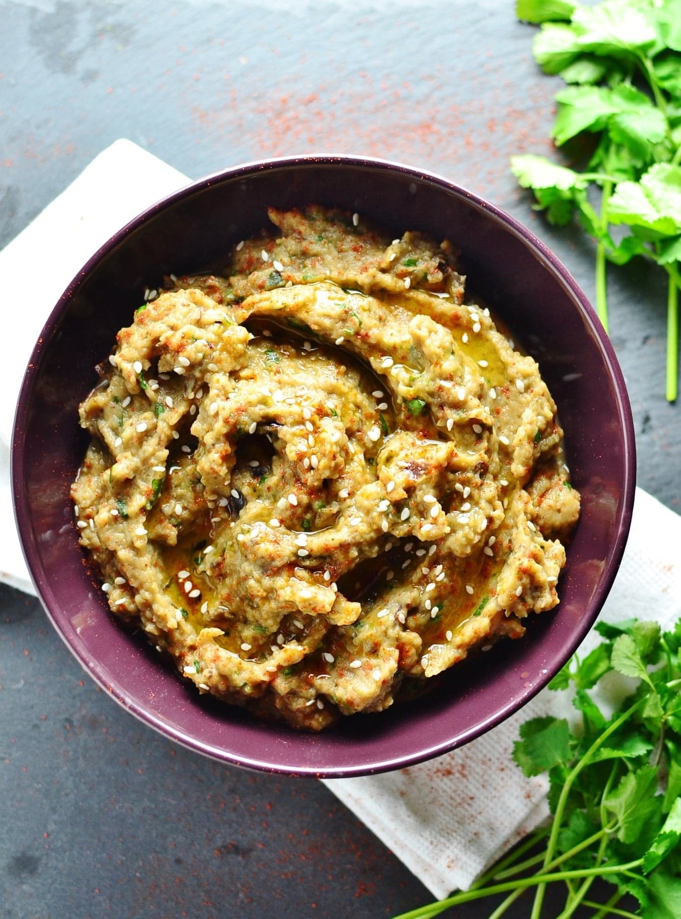 Eggplant dip in purple bowl on top of white cloth, with fresh cilantro in top and bottom right corners.