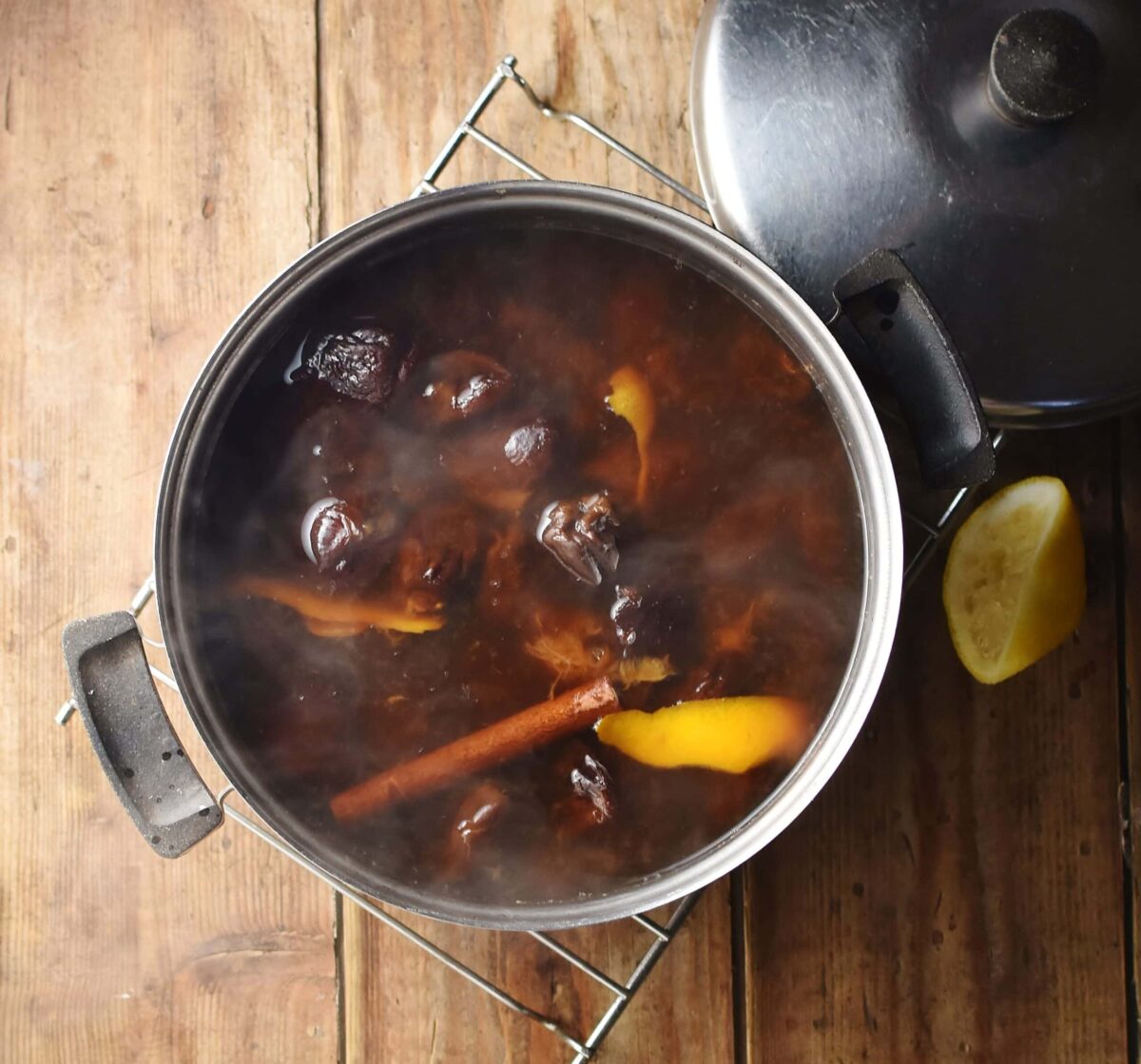 Cooked pruned, orange rind and cinnamon stick and water in pot with steam visible, lemon piece and lid in background.
