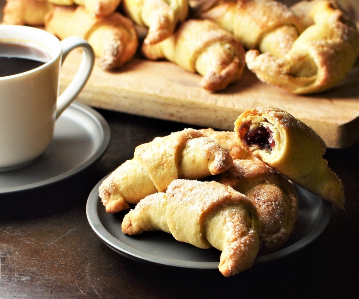Side view of rogaliki on top of saucer and wooden board and white cup with coffee to the left.