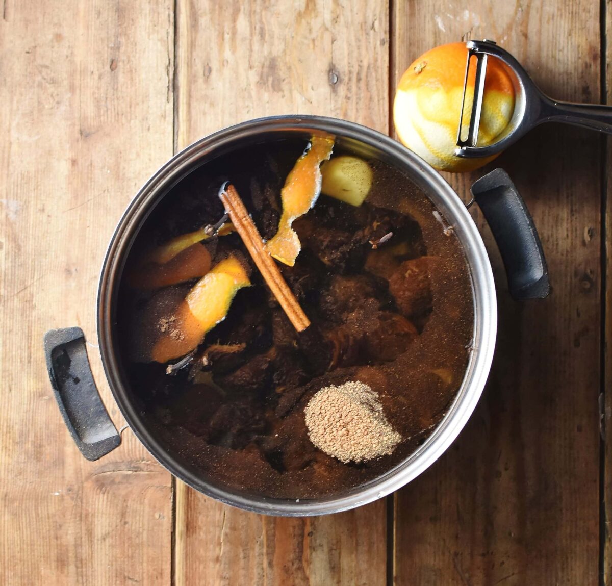 Prunes, orange rind, cinnamon stick and spices with water in pot, with orange and vegetable peeler in background.