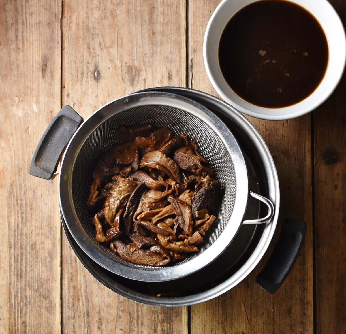 Mushrooms in strainer over pot, with brown liquid in white bowl in top right corner.