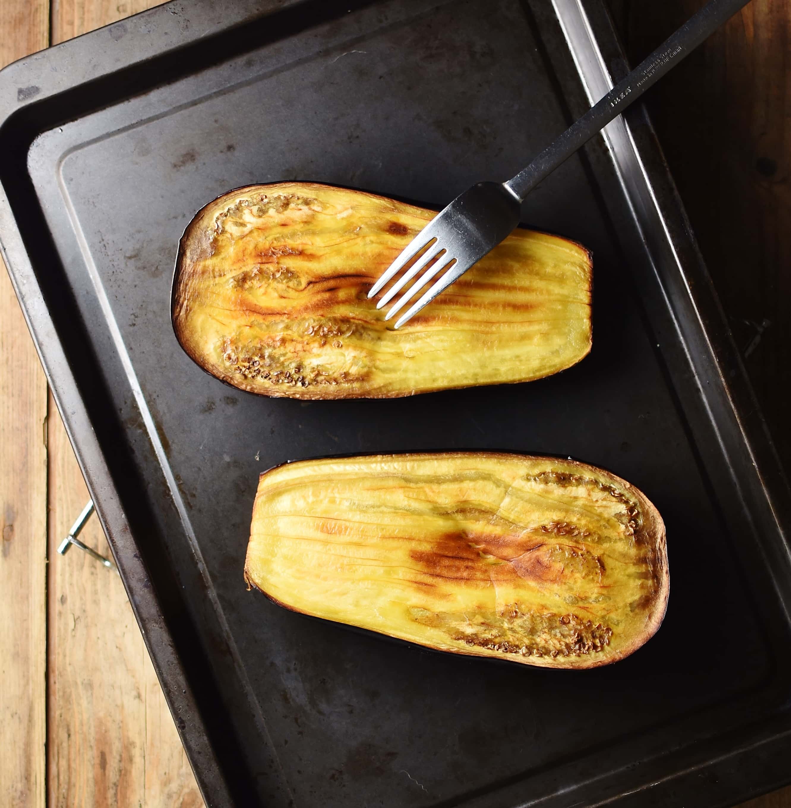 Roasted eggplant halves with fork on top of dark tray.