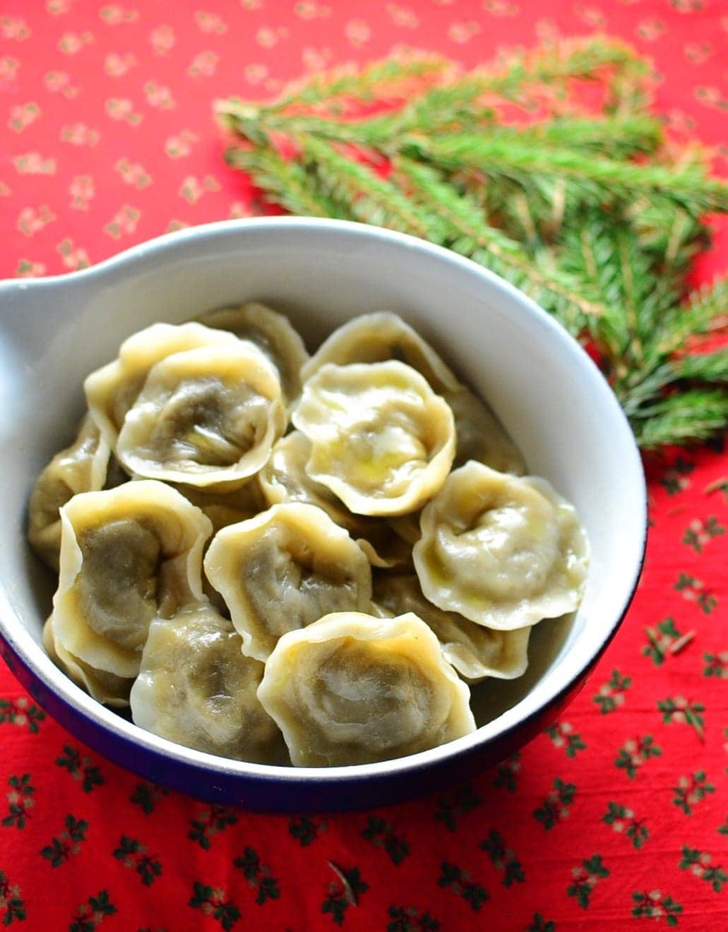 Polish Christmas Eve porcini dumplings (uszka) in white bowl with pine tree branch on red cloth with hollies pattern.