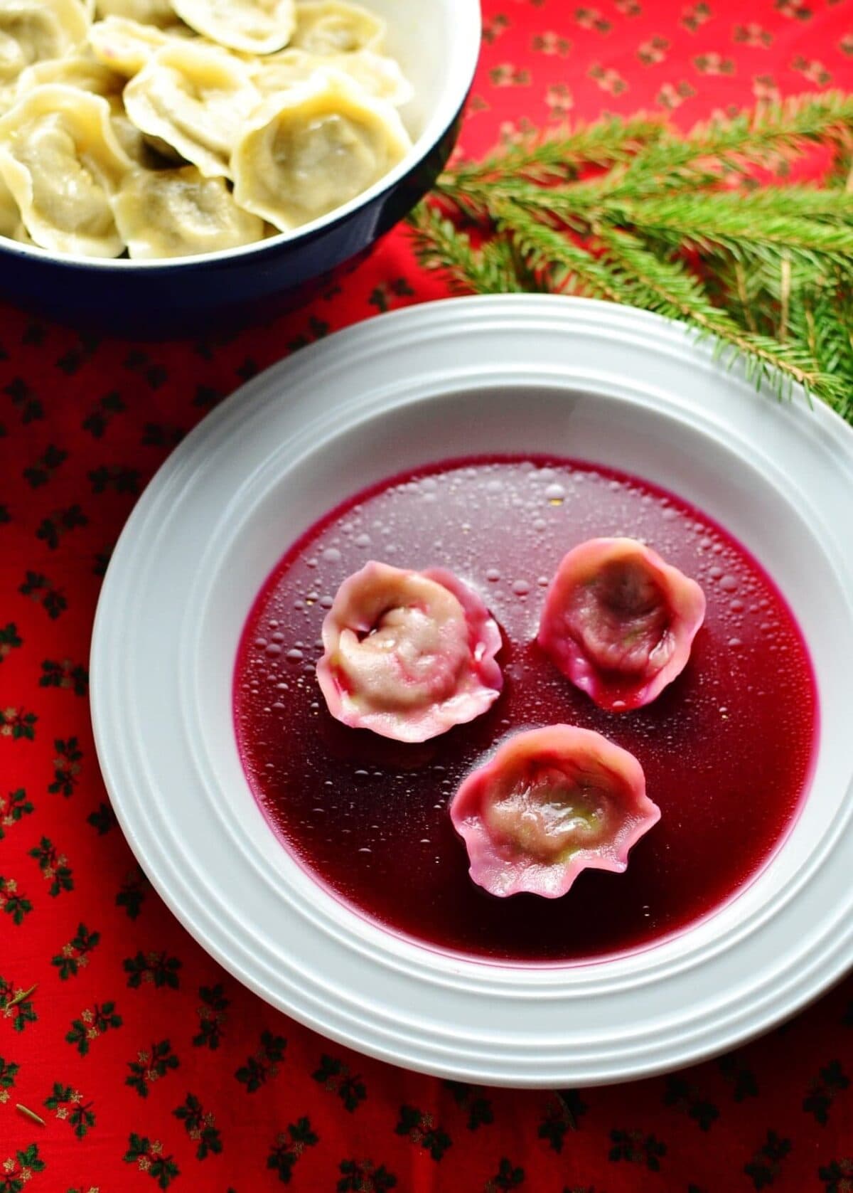 Polish borscht with Christmas uszka dumplings on white plate with uszka in bowl and pine tree branch on red cloth.