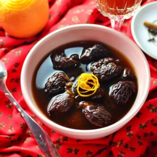 Dried fruit compote with orange rind in white bowl, with orange juice in glass and spoon in background on top of red cloth with Christmas pattern.