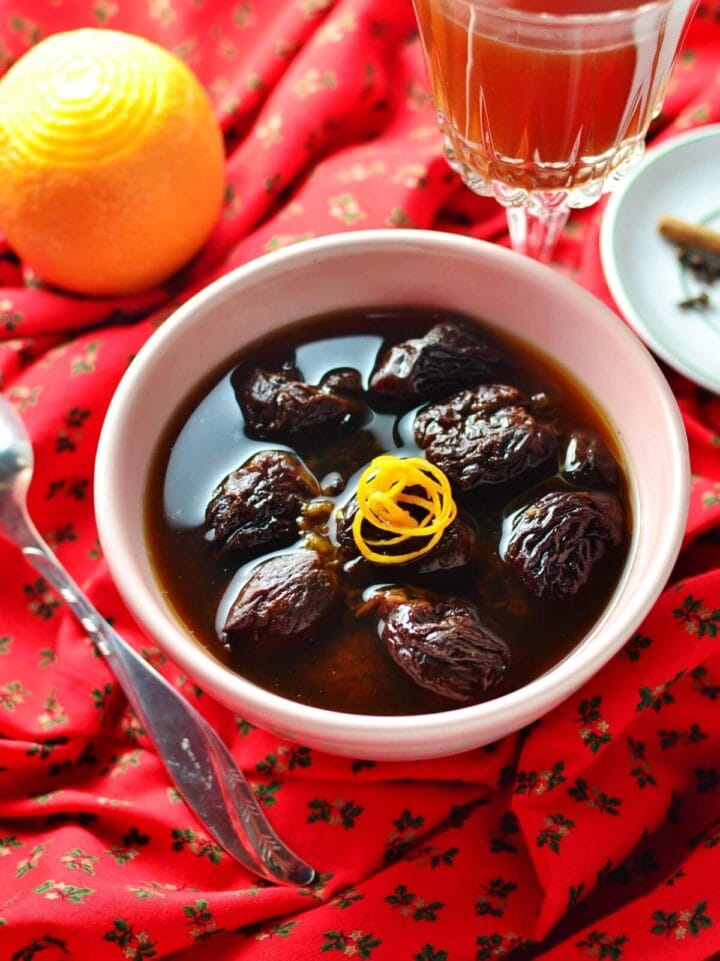 Dried fruit compote with orange rind in white bowl, with orange juice in glass and spoon in background on top of red cloth with Christmas pattern.