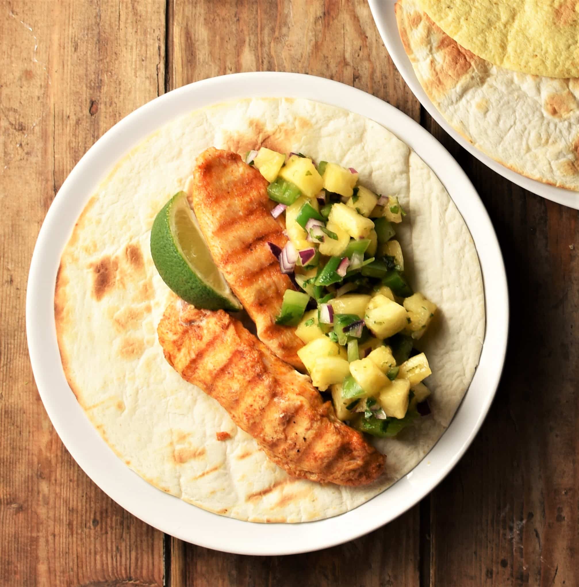 Open tortilla with grilled chicken, pineapple salsa and lime on plate.