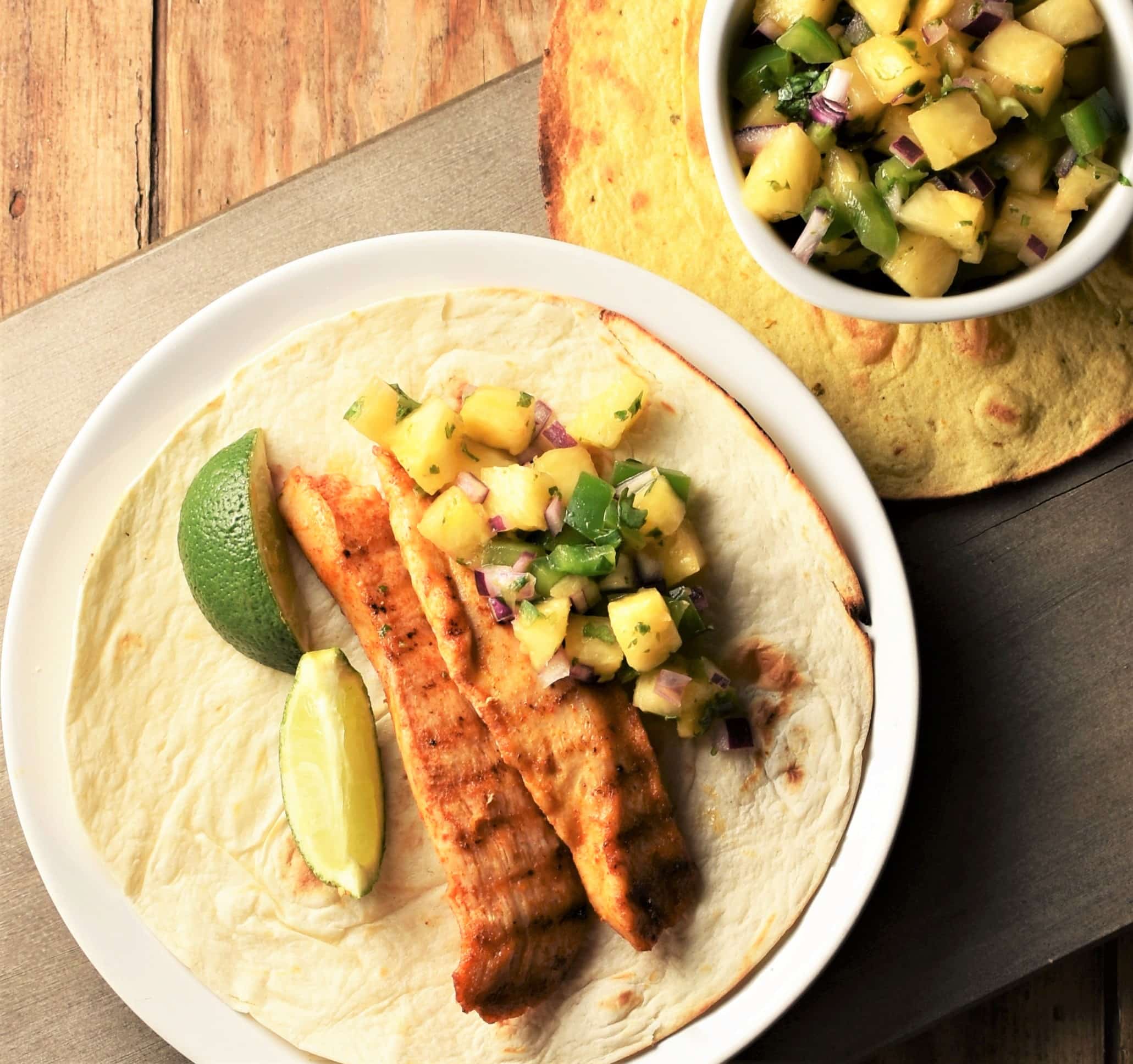 2 strips of grilled chicken with lime wedges and pineapple salsa in open tortilla with salsa in top right corner.