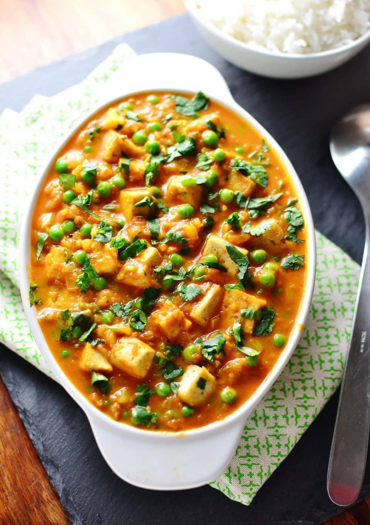 Top down view of tofu curry with peas in white oval dish on top of green-and-white cloth, with large spoon to right and cooked rice in white bowl in top right corner.