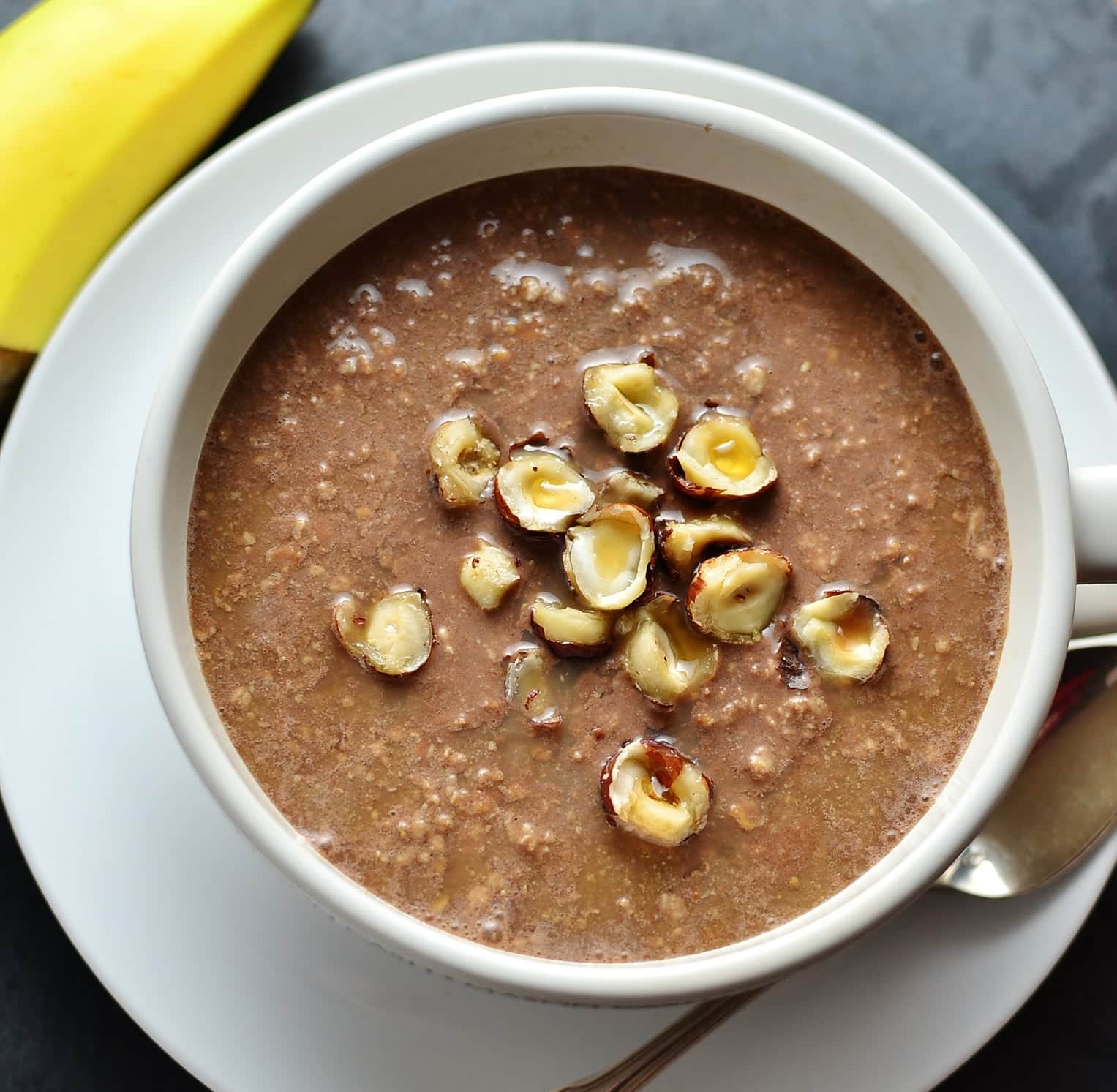 Top down view of chocolate overnight oats topped with crushed hazelnuts in white cup on top of white saucer with spoon and banana in background.