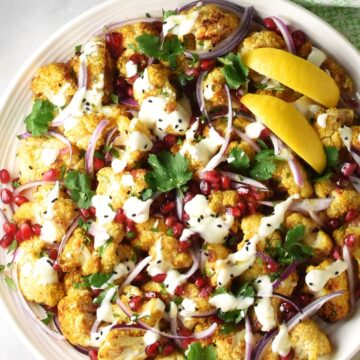 Top down close-up view of curried cauliflower salad with creamy dressing and lemon wedges on large plate.